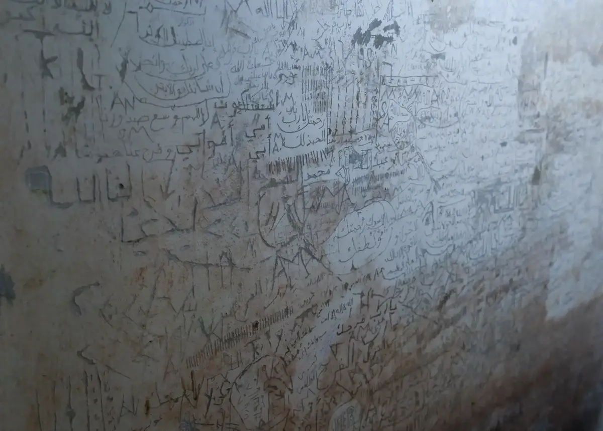 A close-up of a heavily graffitied wall covered in dense, overlapping Arabic script and crude drawings, with the phrase…