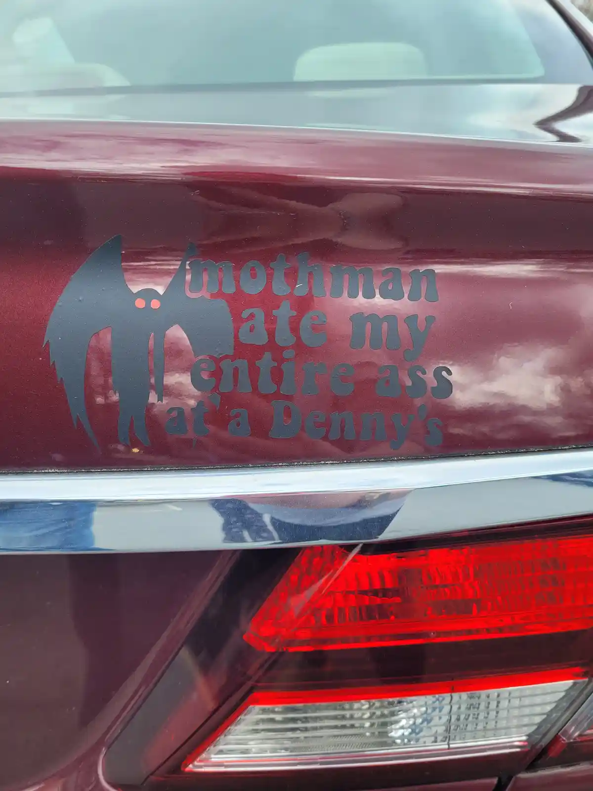 A dark gray vinyl sticker on the rear of a maroon car features a silhouette of the Mothman, a legendary cryptid with…