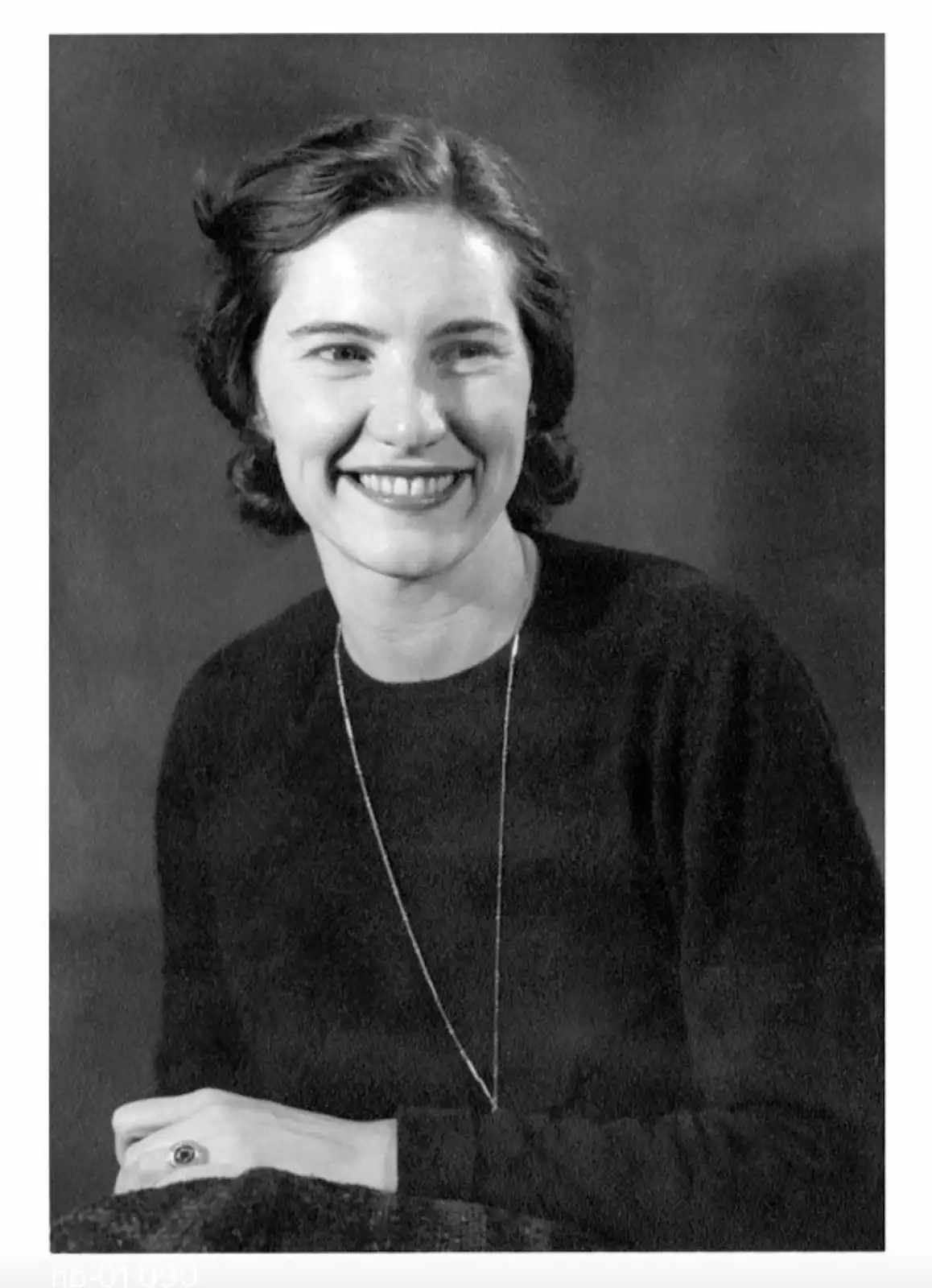 A black-and-white portrait of a smiling woman with short dark hair, wearing a dark sweater and a thin necklace, her hand…