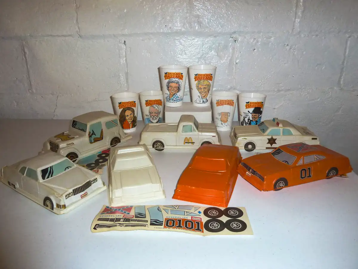 A collection of vintage The Dukes of Hazzard McDonald's Happy Meal toys and cups are displayed on a white surface against a…