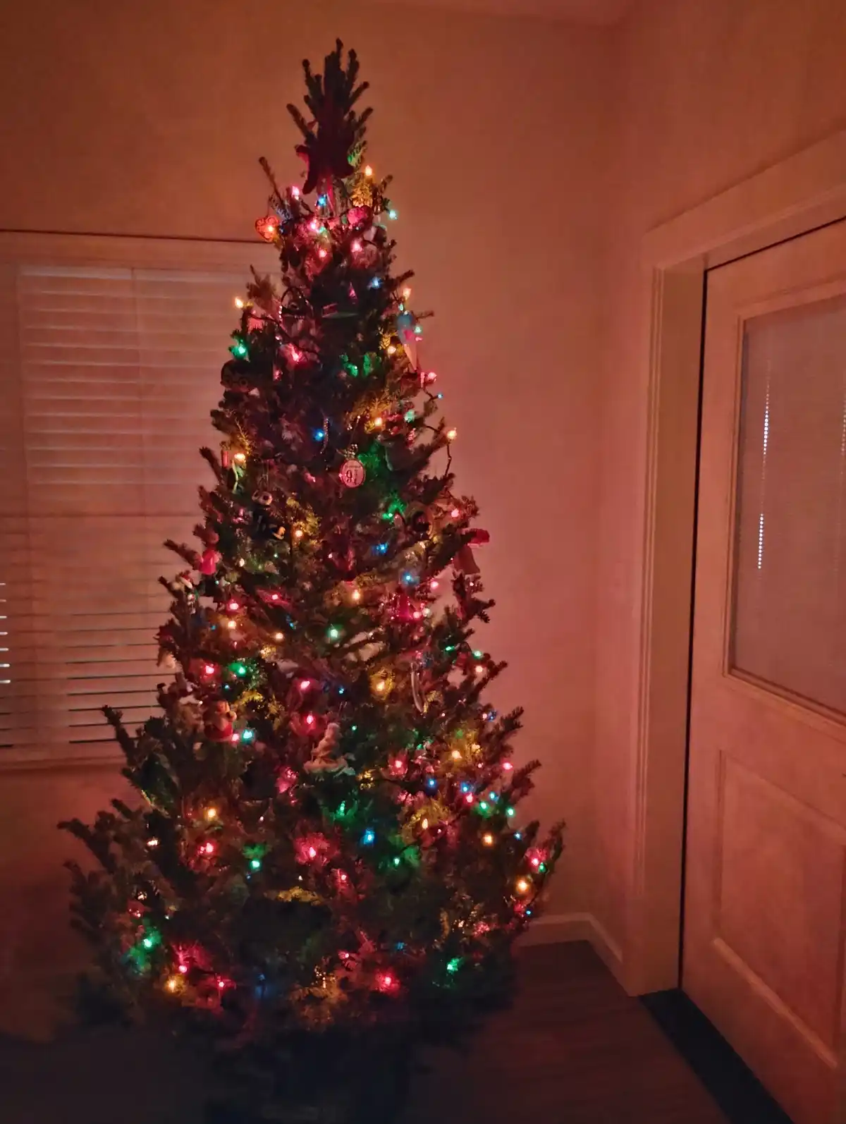 A brightly lit, densely decorated artificial Christmas tree stands in a dimly lit room, adorned with multicolored lights and…