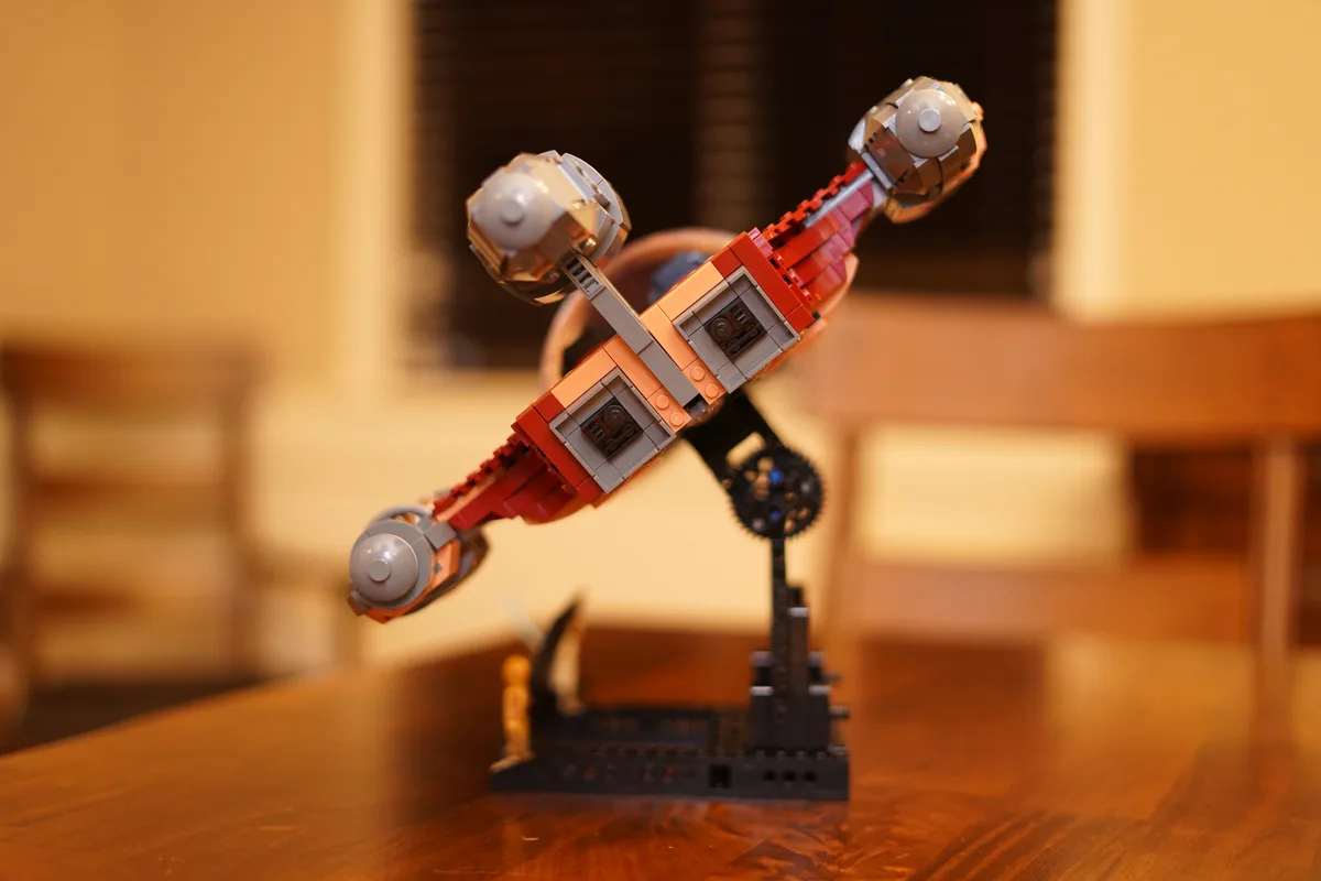 A red and gray Lego Star Wars ship model is balanced precariously on a black Lego stand on a wooden table, appearing to…