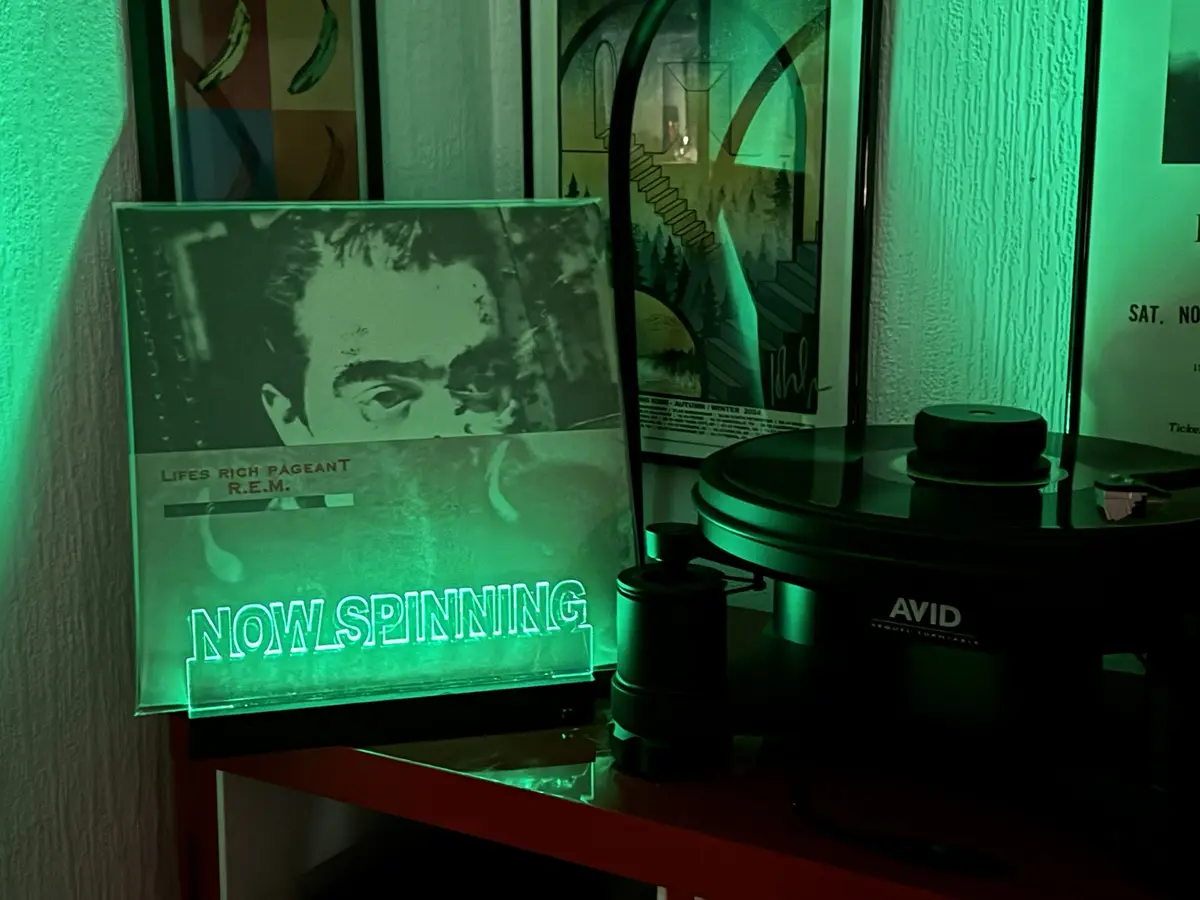 A green-lit vinyl record player sits next to a glowing album cover for R.E.M.'s 'Lifes Rich Pageant', with the text 'NOW…