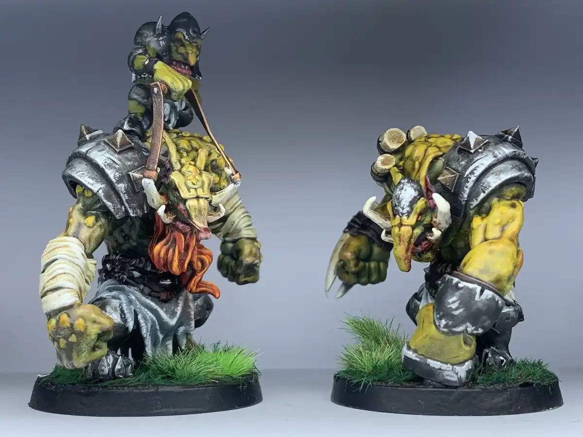 Two painted fantasy miniatures of green-skinned, horned, armored ogres stand side by side on grassy bases against a gray…