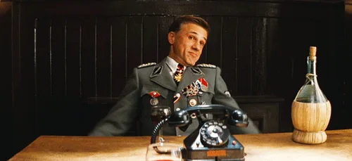 A man in a decorated military uniform sits at a wooden desk with a rotary phone and a bottle, progressively spreading his…