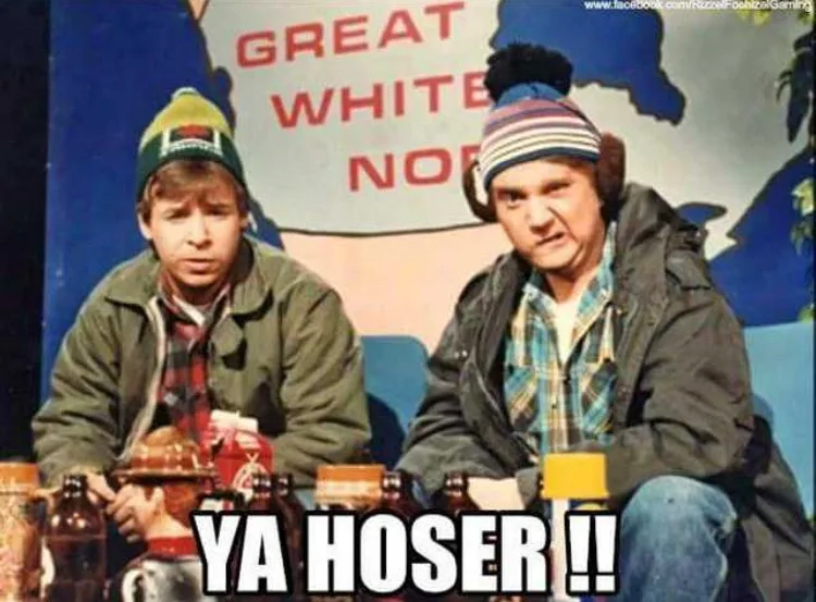 Two men in winter hats and jackets sit at a table with beer bottles, in front of a backdrop reading 'GREAT WHITE NORTH'. The…