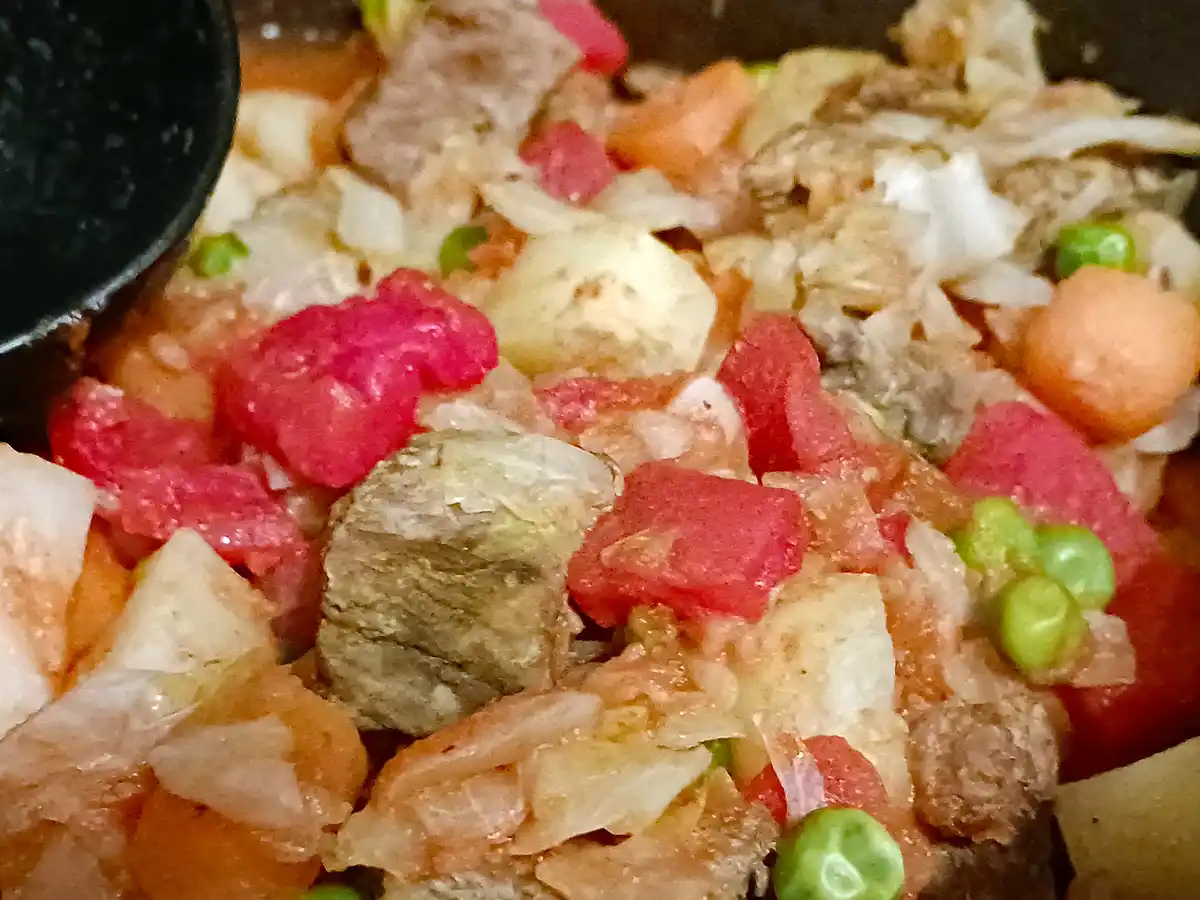 A close-up photo of a stew or curry with chunks of meat, diced tomatoes, onions, peas, and carrots in a dark pot, with a…