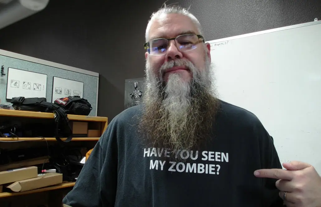 A man with a long gray beard and glasses wears a black t-shirt that reads 'HAVE YOU SEEN MY ZOMBIE?' and points to the text…