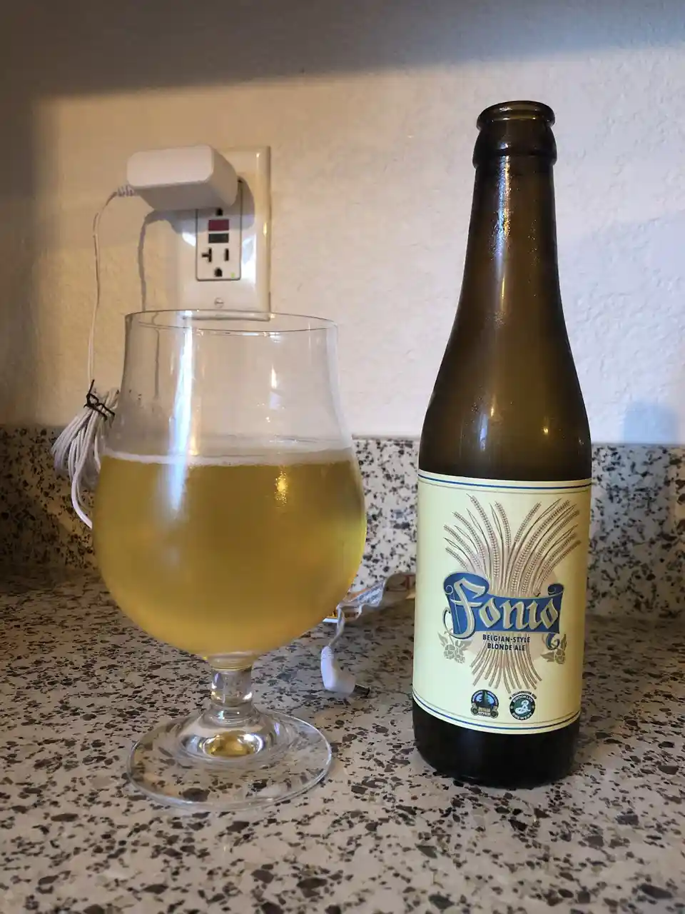 A glass of golden beer sits next to a brown bottle labeled 'Fonio Belgian-Style Blonde Ale' on a speckled countertop, with a…