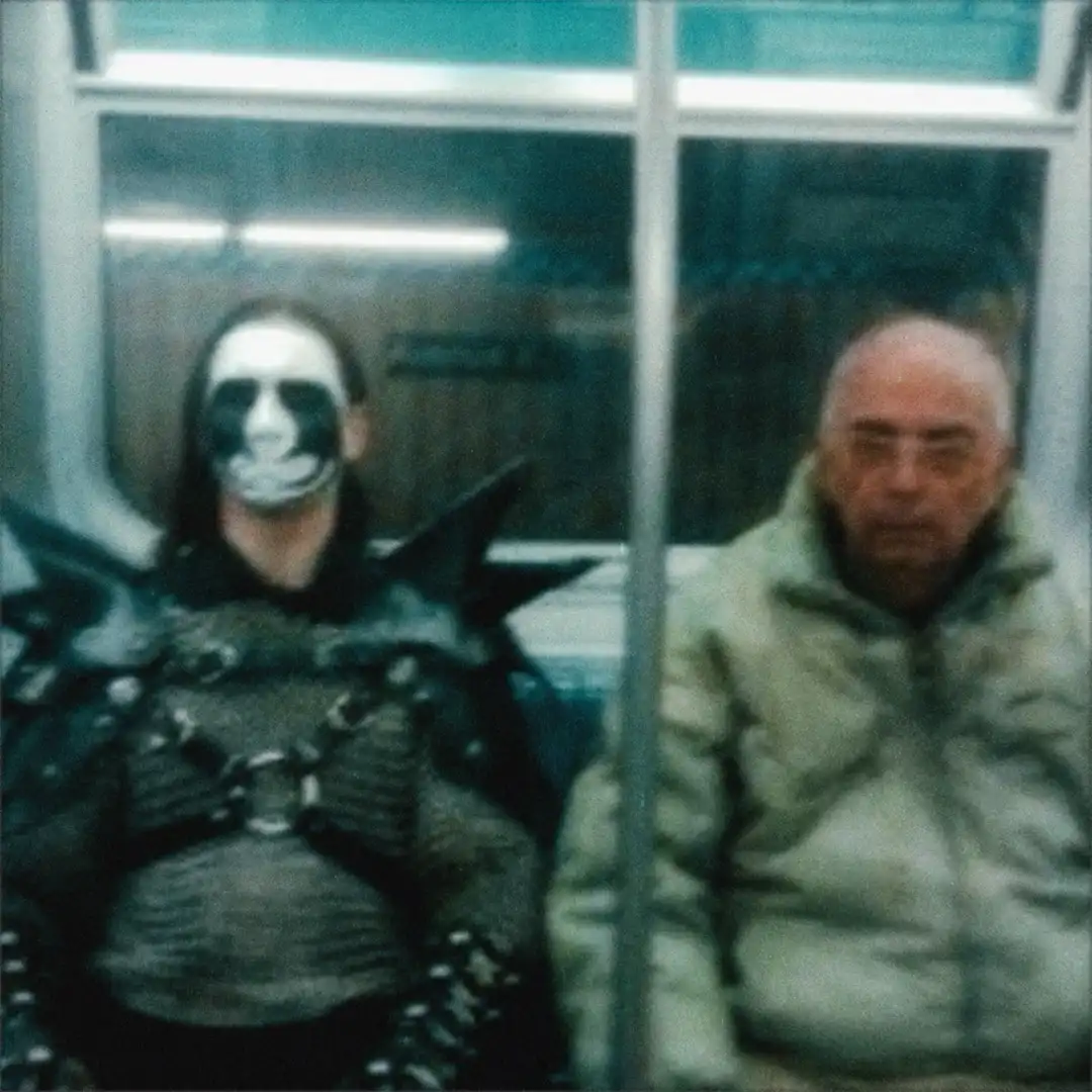 Two people sit side by side on a subway train. The person on the left wears a full-face skull makeup and dark, spiky costume…