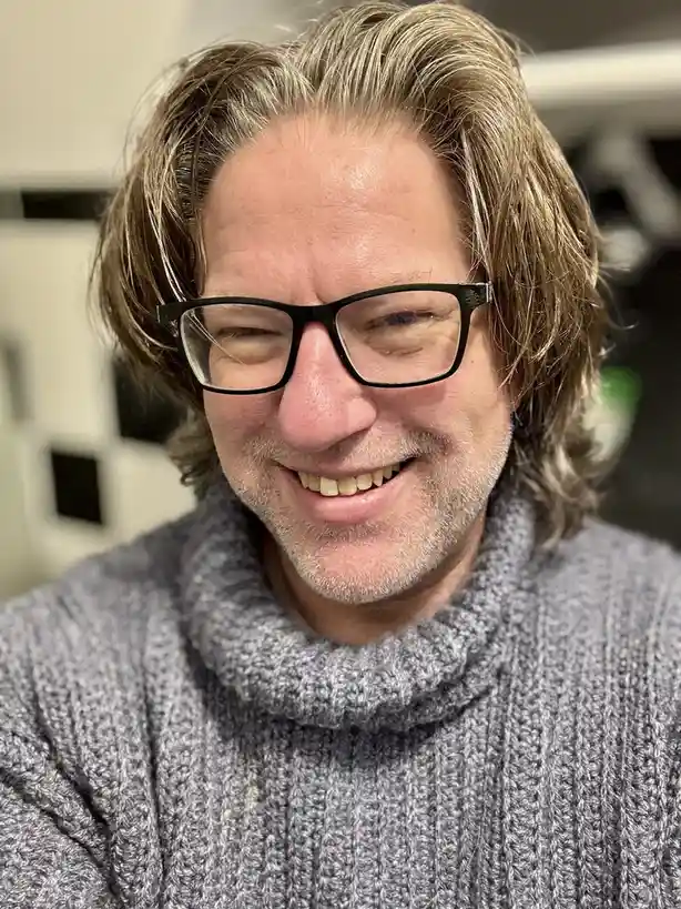 A close-up photo of a middle-aged man with shoulder-length, wavy, salt-and-pepper hair, wearing black-rimmed glasses and a…
