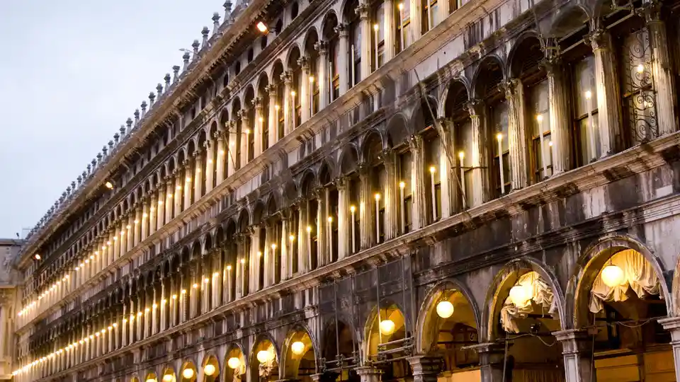 Golden light dances across ornate arches and the intricately carved facade of a grand Venetian building, its Byzantine…