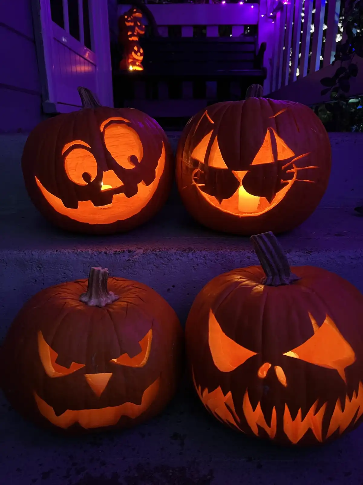 Four carved pumpkins with glowing candles inside sit on concrete steps at night, illuminated by purple ambient lighting. The…