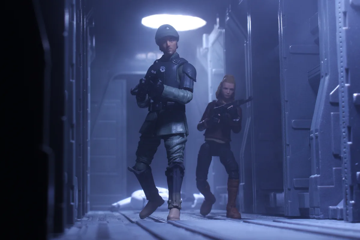 Two Star Wars action figures, one male and one female in combat gear, stand in a dimly lit, foggy spaceship corridor with a…