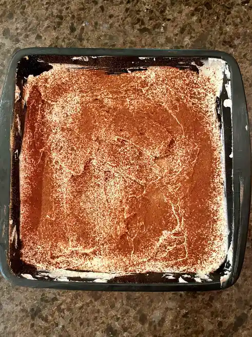 A top-down photo shows a square black baking dish containing a dessert covered in cocoa powder, with the powder arranged to…