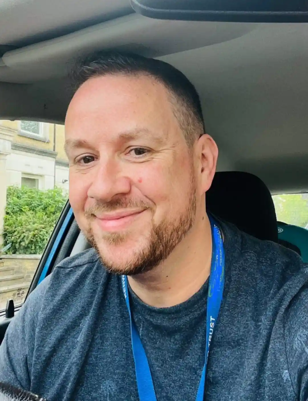 a man with a beard and short hair smiles while sitting in a car, wearing a blue lanyard around his neck. The lanyard has…