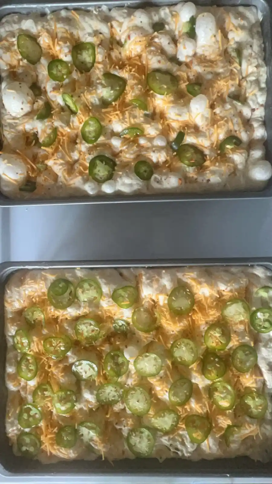 Two identical metal baking trays hold the same unbaked dish, topped with shredded cheese and sliced jalapeños. The top tray…