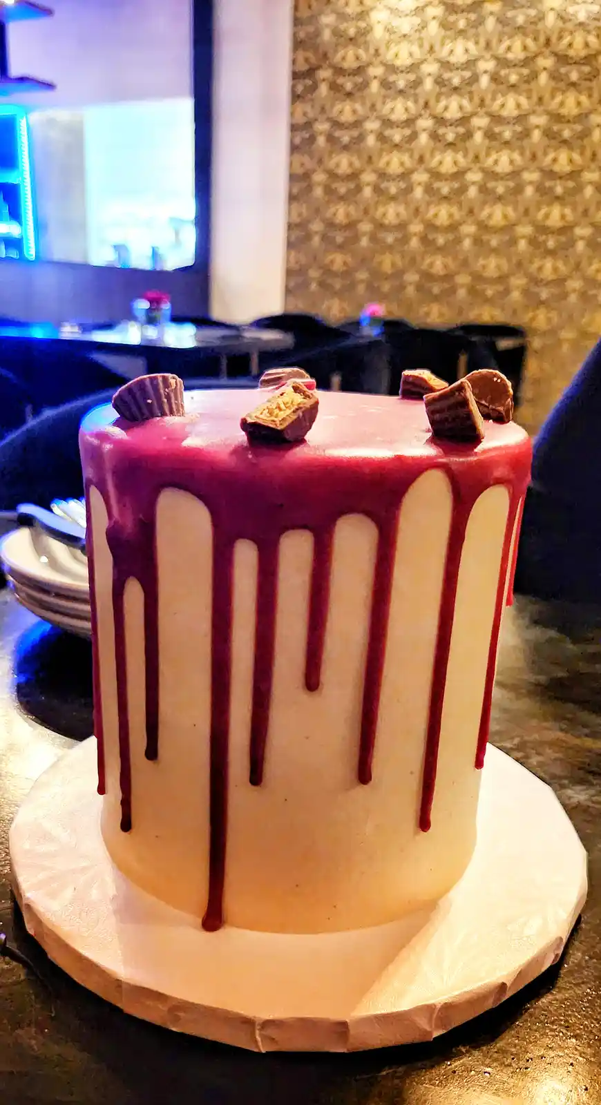 A cylindrical cake with white frosting and red icing dripping down its sides, topped with broken peanut butter cups, sits on…