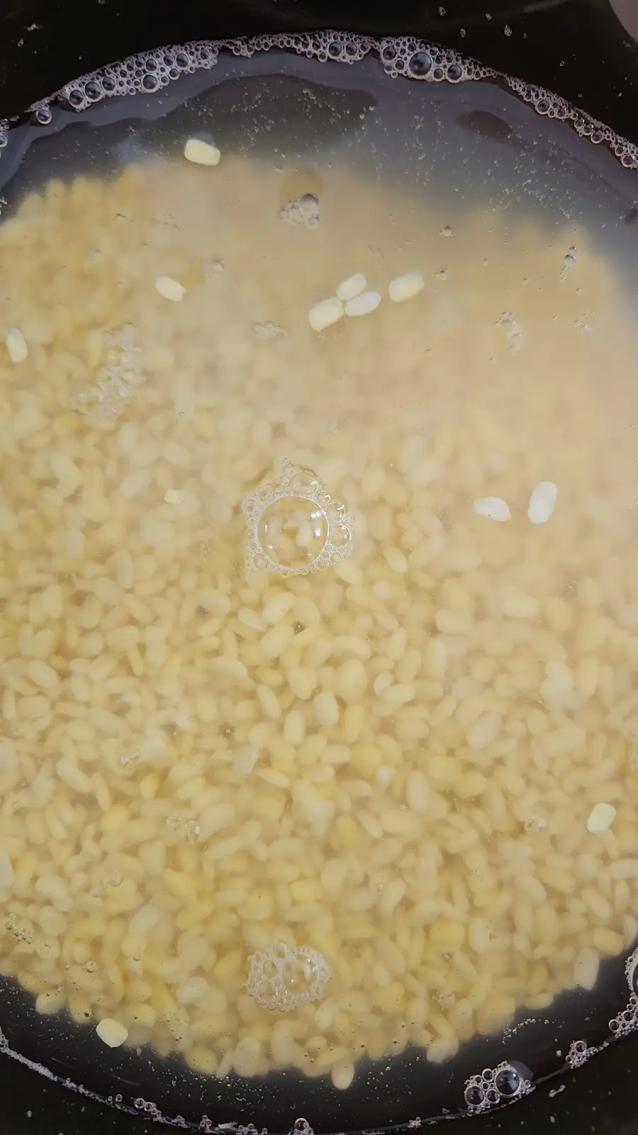 A close-up photo shows small, pale yellow rice grains boiling in water inside a dark pan, with bubbles forming on the…