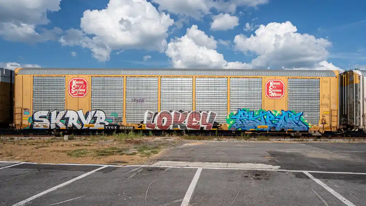A yellow Kansas City Southern Lines freight train car sits on tracks under a blue sky with white clouds, covered in colorful…
