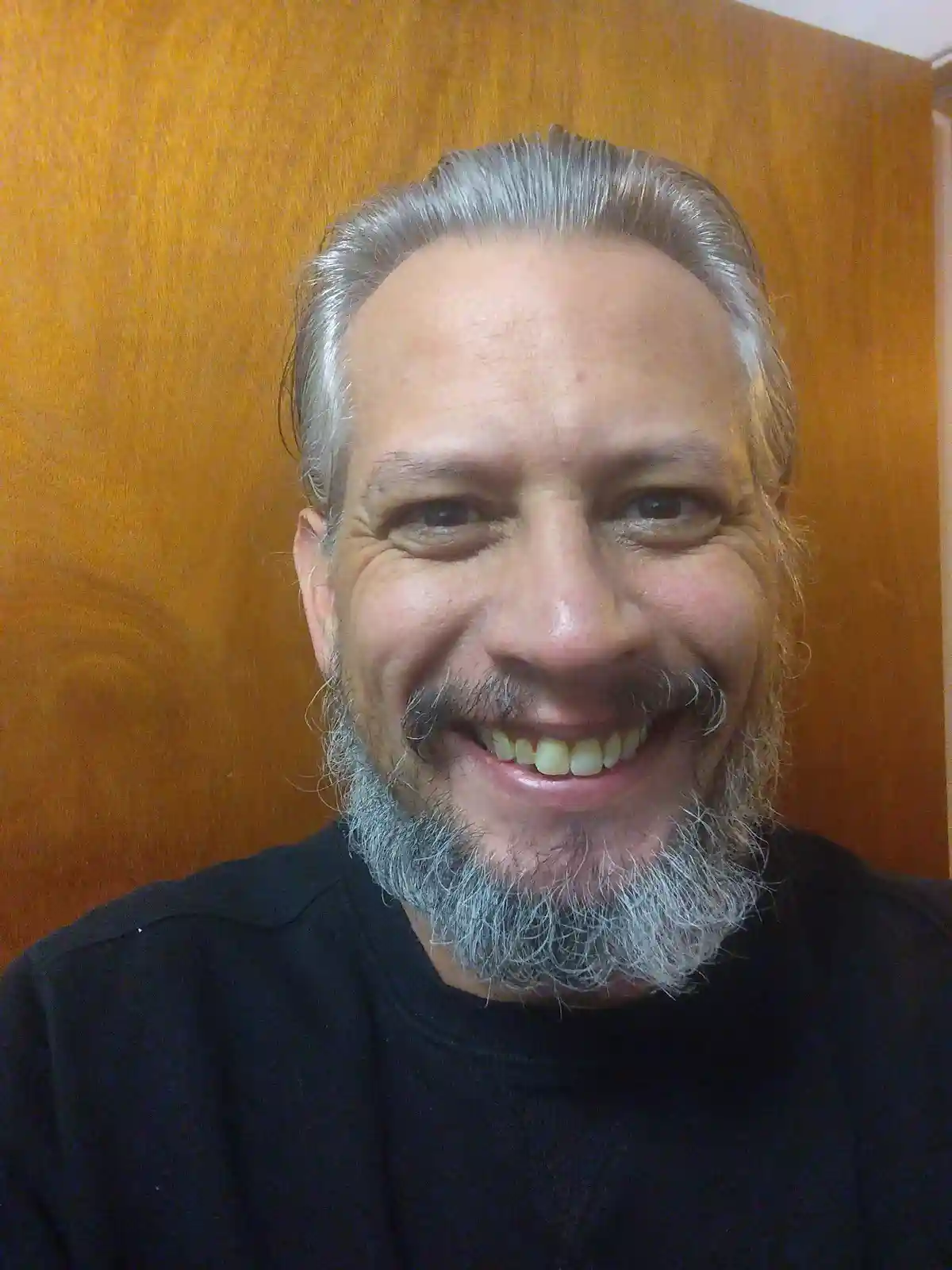 A middle-aged man with gray hair and a gray beard smiles broadly at the camera while wearing a black shirt, standing in…