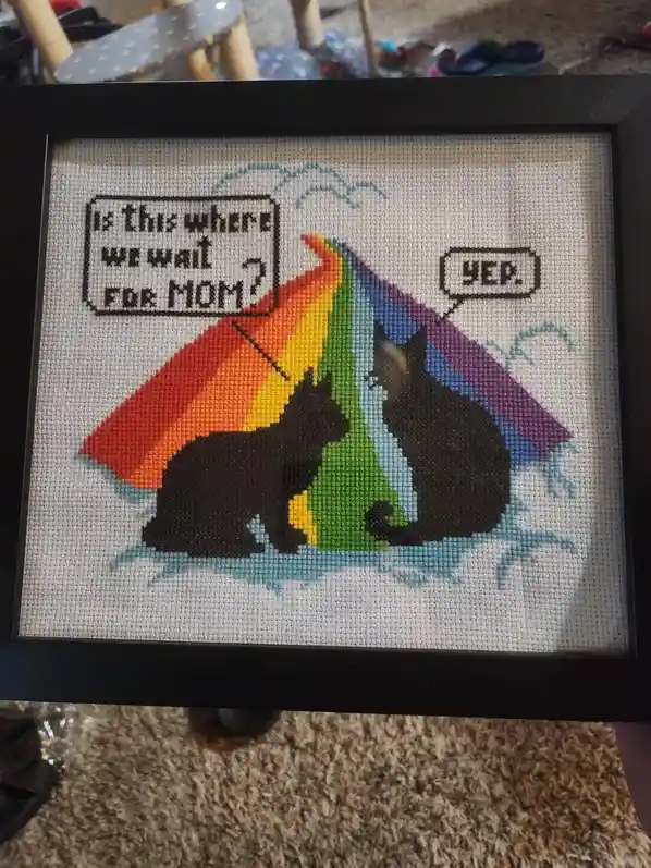 A framed cross-stitch artwork depicts two black cat silhouettes sitting under a rainbow-colored umbrella in the rain. One…