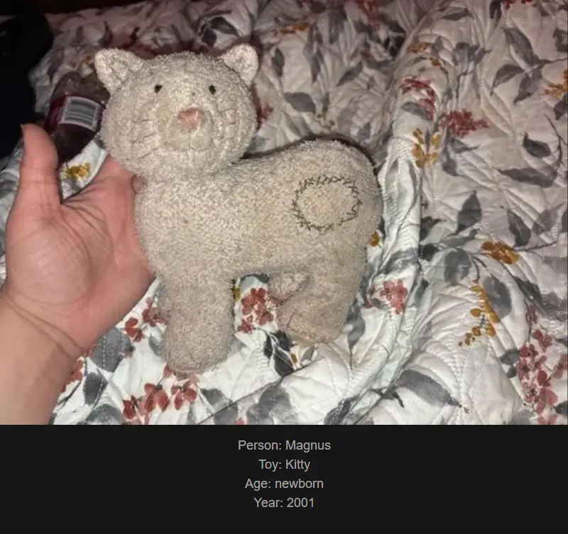 A hand holds a small, beige, fuzzy stuffed cat toy with a stitched spiral on its side, resting on a floral-patterned quilt…