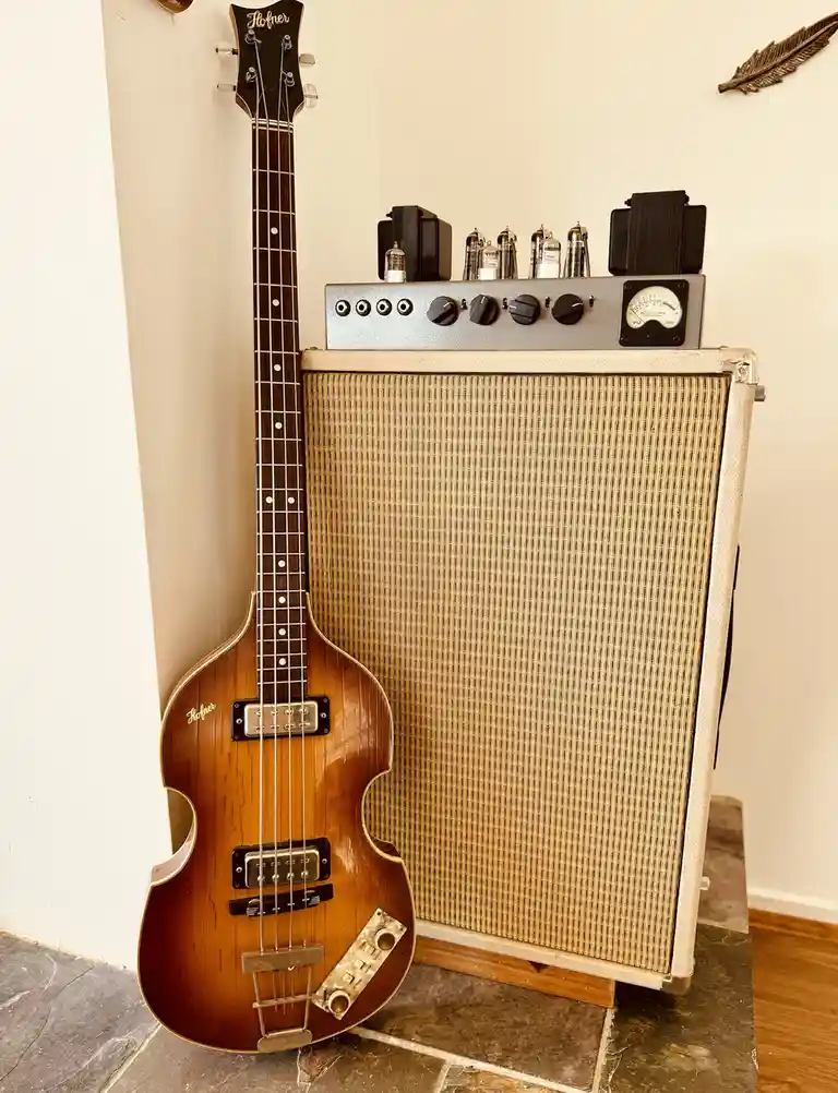 A vintage Hofner bass guitar leans against a vintage tube amplifier with visible vacuum tubes and knobs. The image has…