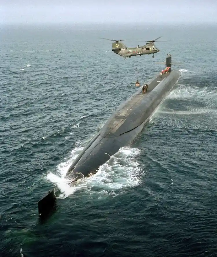 An aerial photo shows a dark gray submarine partially surfaced in choppy blue ocean water, with a military helicopter…