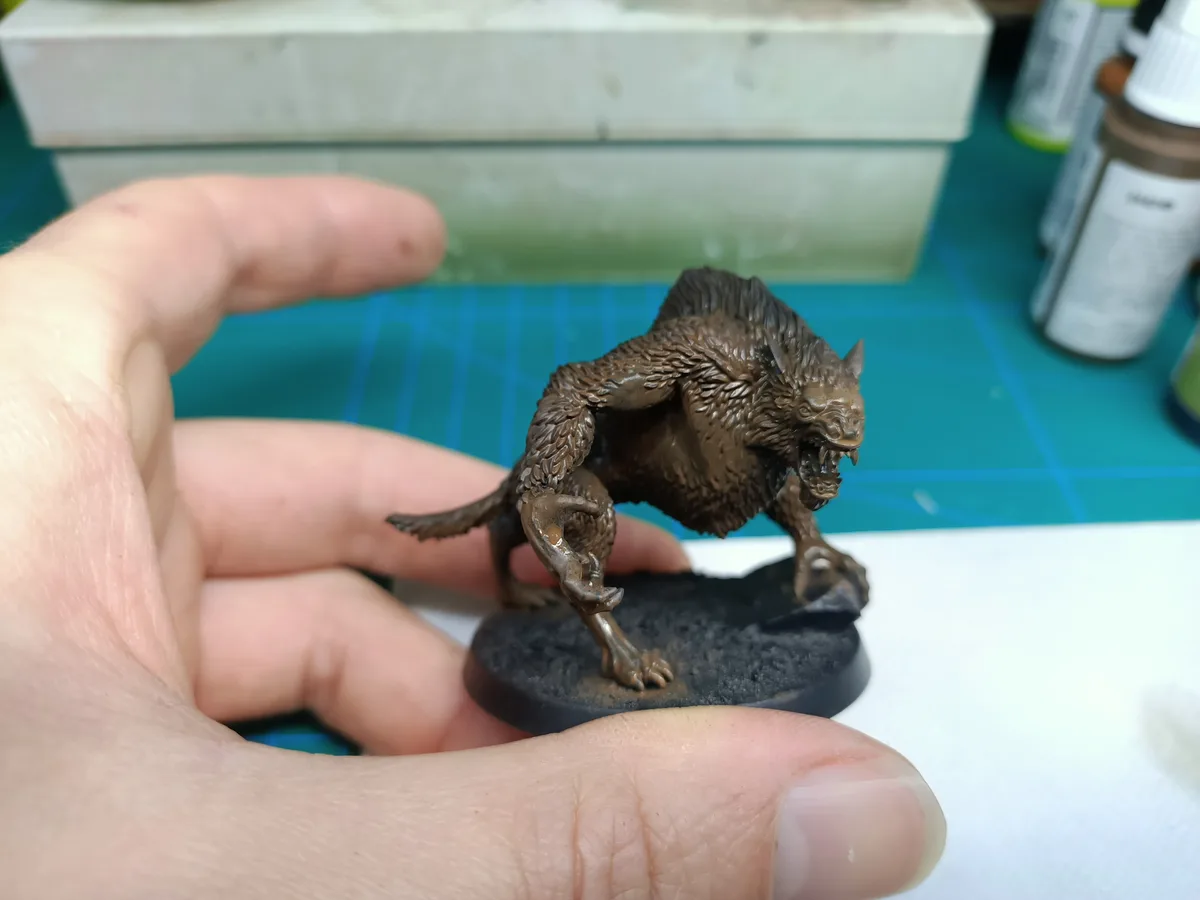 A person holds a small, unpainted brown miniature of a furry, snarling beast with sharp teeth and claws, standing on a black…
