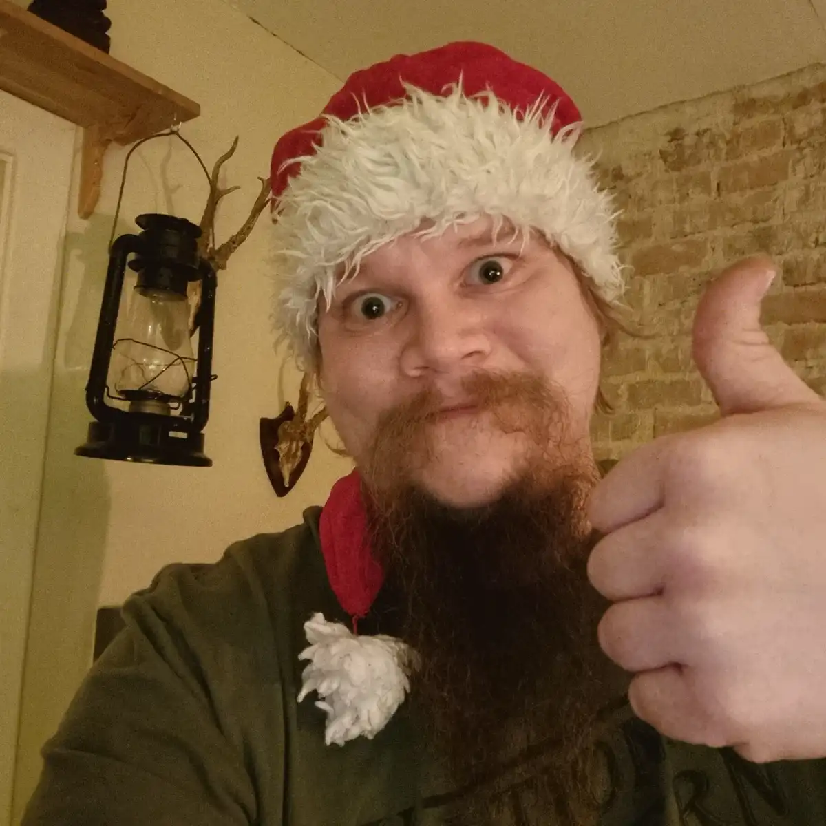 A man with a long beard and mustache wears a red and white Santa hat and gives a thumbs-up while making a wide-eyed…