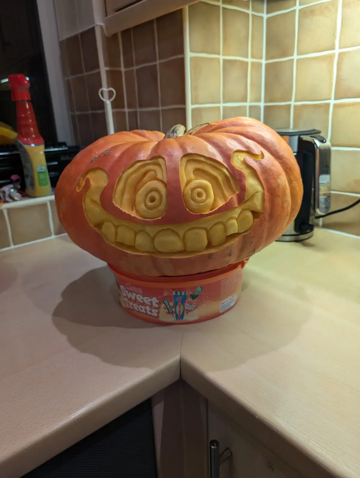 A large orange pumpkin with a carved jack-o'-lantern face, featuring spiral eyes and a toothy grin, sits atop a container of…