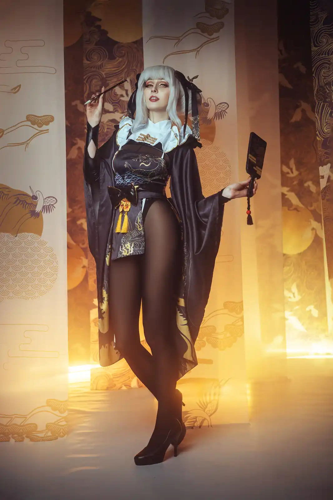 A person in a stylized, modernized Japanese-inspired costume with white hair, holding a brush and a small black fan, poses…