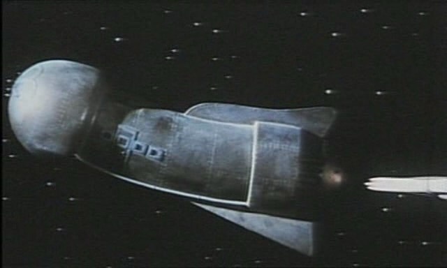 A grainy, low-resolution image shows a silver, cylindrical spaceship with a rounded front, floating in a starfield. The ship…