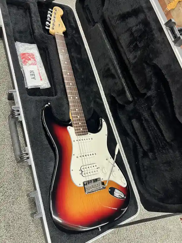 A sunburst Fender Stratocaster electric guitar rests in its open black-lined hard case, with a small plastic-wrapped item…