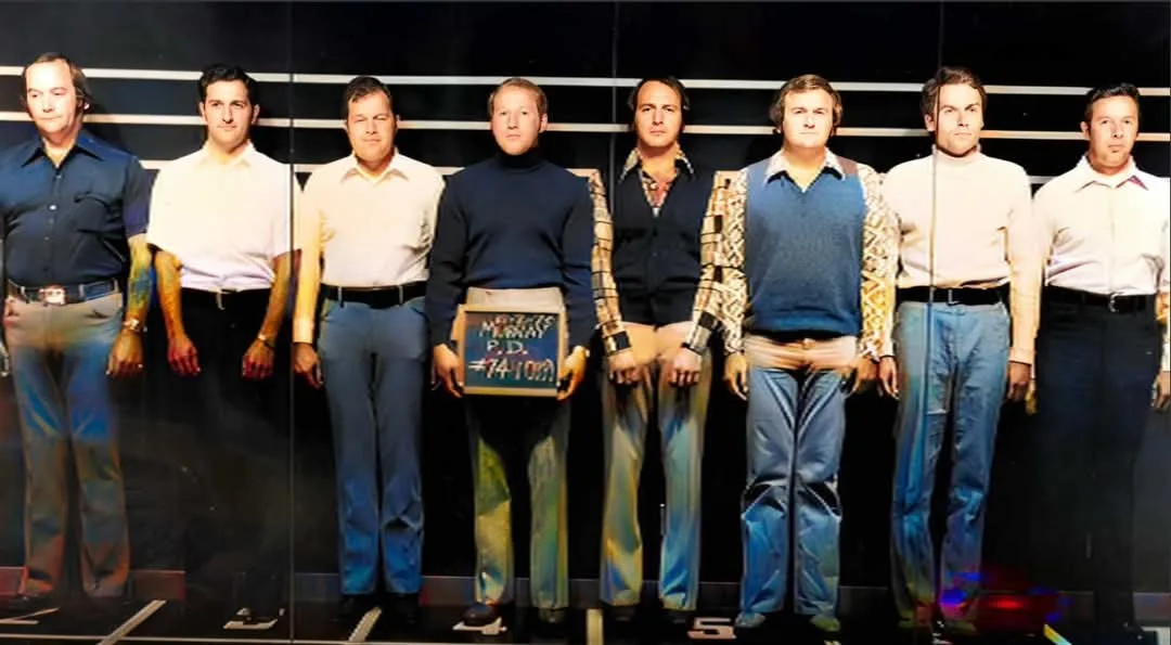 Seven men in 1970s-style clothing stand in a police lineup against a dark background with horizontal white stripes. The man…