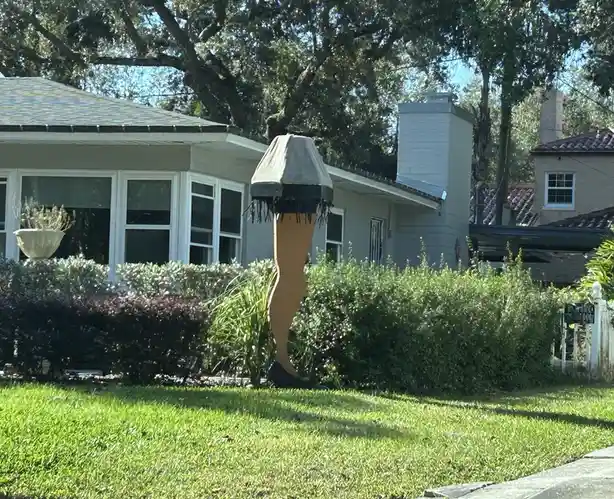 A large, life-sized statue of a leg wearing a black high-heeled shoe and a fringed lampshade on top stands in a grassy front…