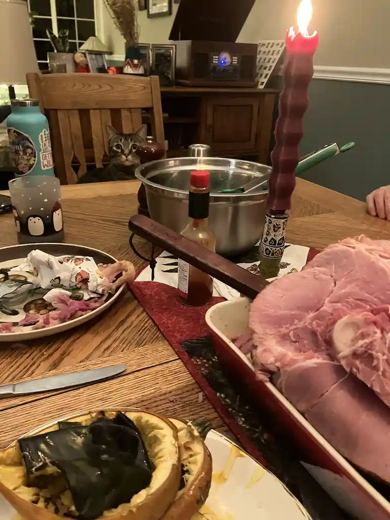 A dining table set for a holiday meal with a large ham in a red dish, a lit purple candle, and a plate with leftover food. A…