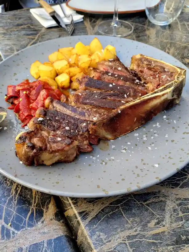 A plate of food features a large, sliced tomahawk steak with a prominent bone, accompanied by roasted potatoes and red…