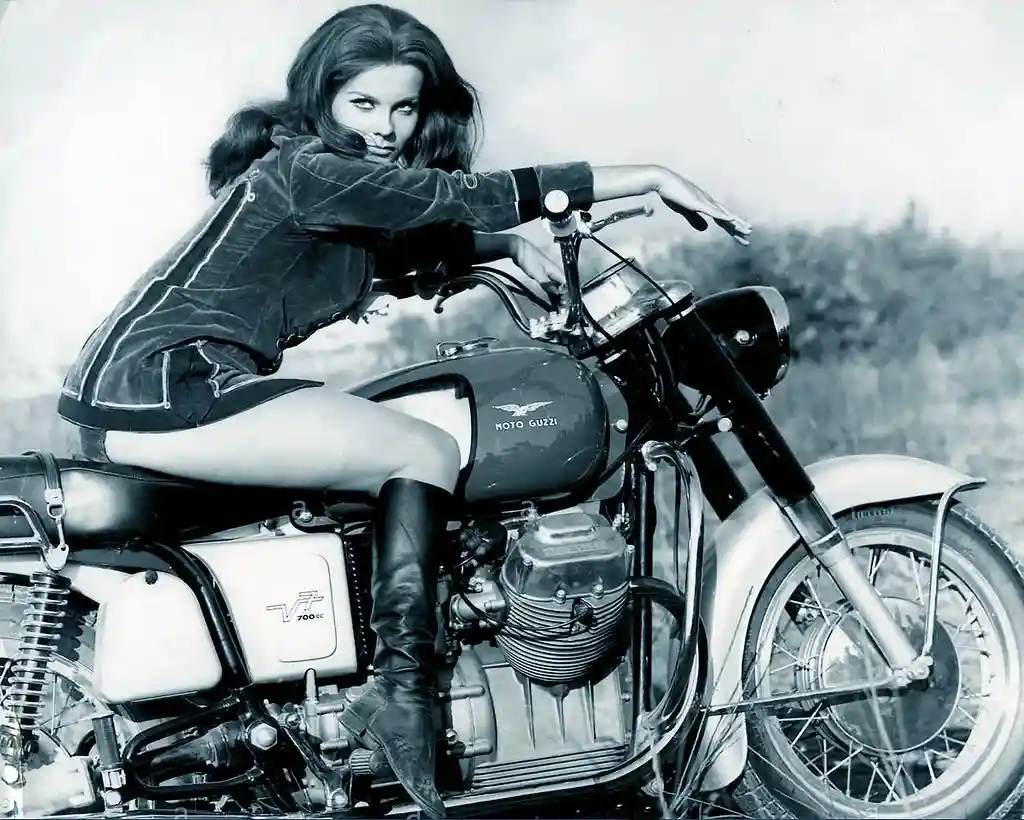 A black-and-white photo shows a woman in a leather jacket and boots sitting on a Moto Guzzi motorcycle, looking directly at…