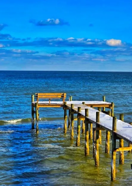 A wooden pier extends into blue ocean water under a bright blue sky with scattered clouds. The pier's surface and…