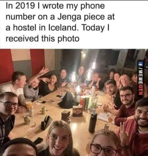 A group of people are gathered around a wooden table in a dimly lit room, smiling and waving at the camera. The table is…