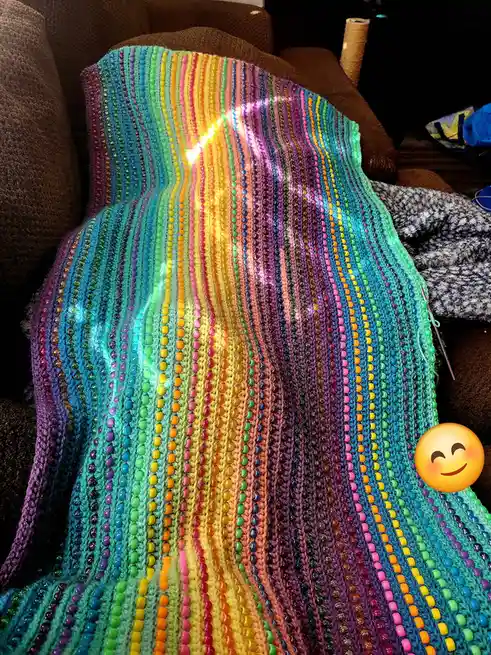 A colorful, hand-crocheted blanket with rainbow stripes of beads is draped over a brown couch, illuminated by a bright light…