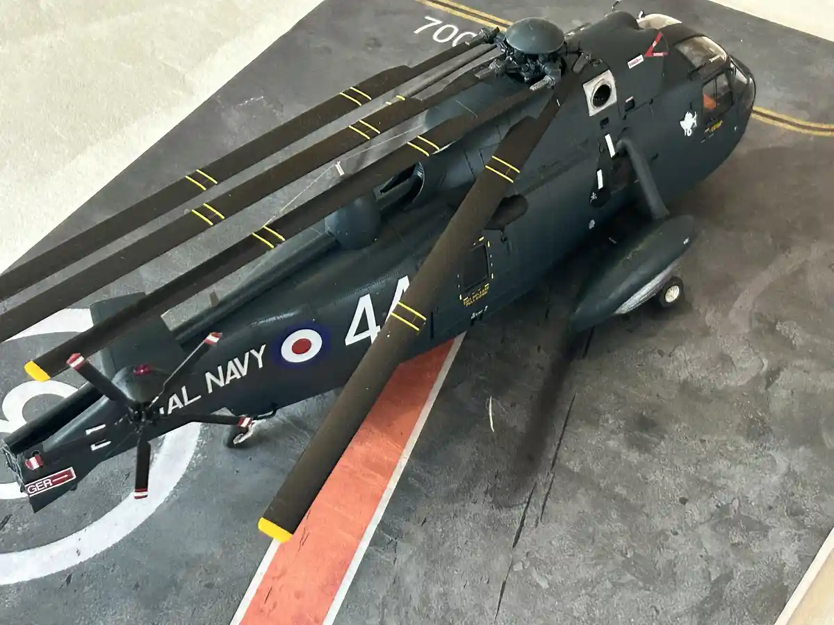 An overhead photo of a dark green Royal Navy helicopter model on a grey deck with orange and white markings. The helicopter…