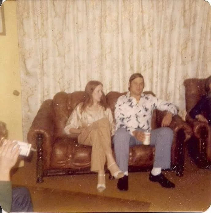 A grainy, vintage photo shows a man and woman sitting on a brown leather couch in a dated living room with patterned…