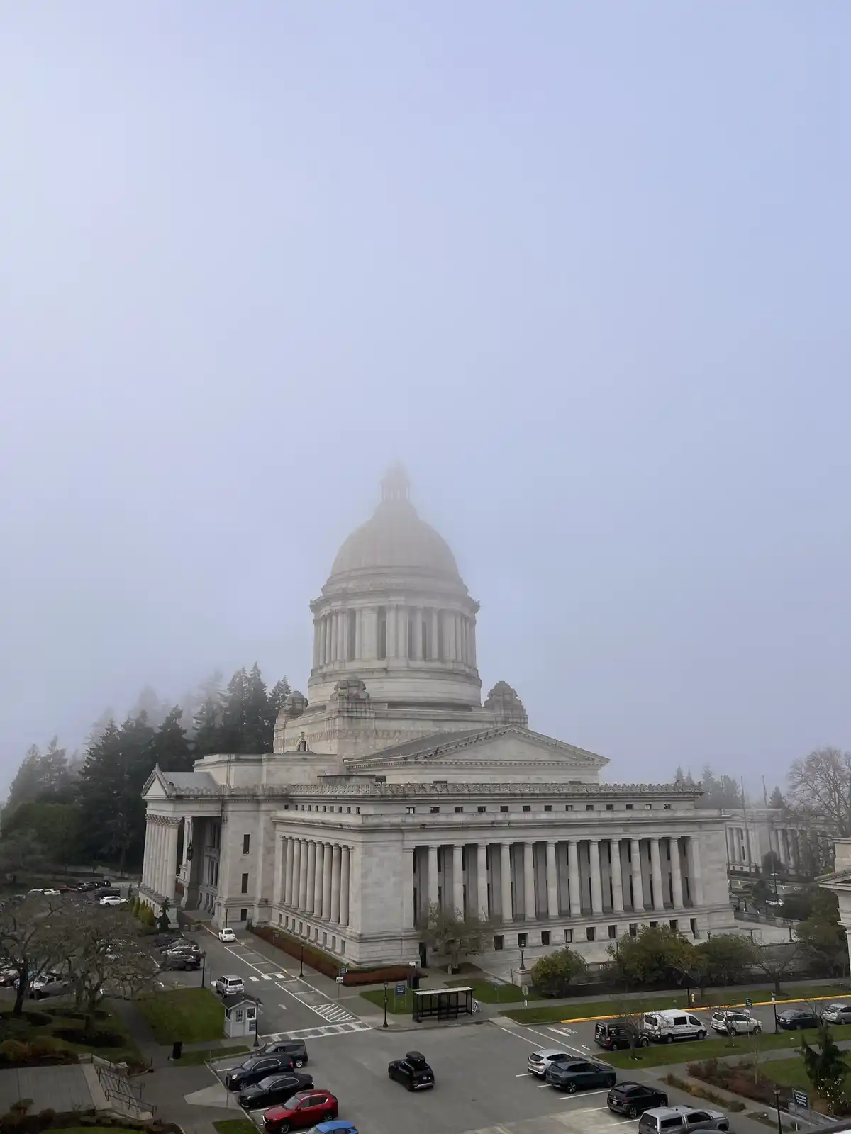 A large, classical-style stone capitol building with a prominent dome is partially obscured by thick fog, with cars parked…
