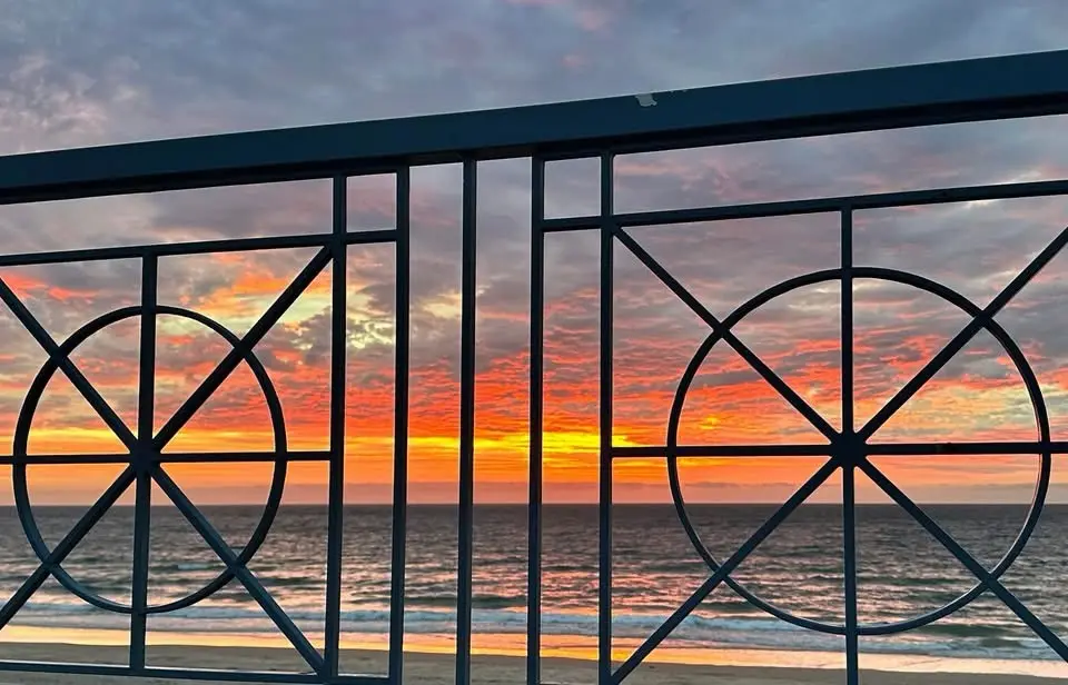 A photograph of a vibrant sunset over the ocean viewed through a dark metal balcony railing with circular and geometric…