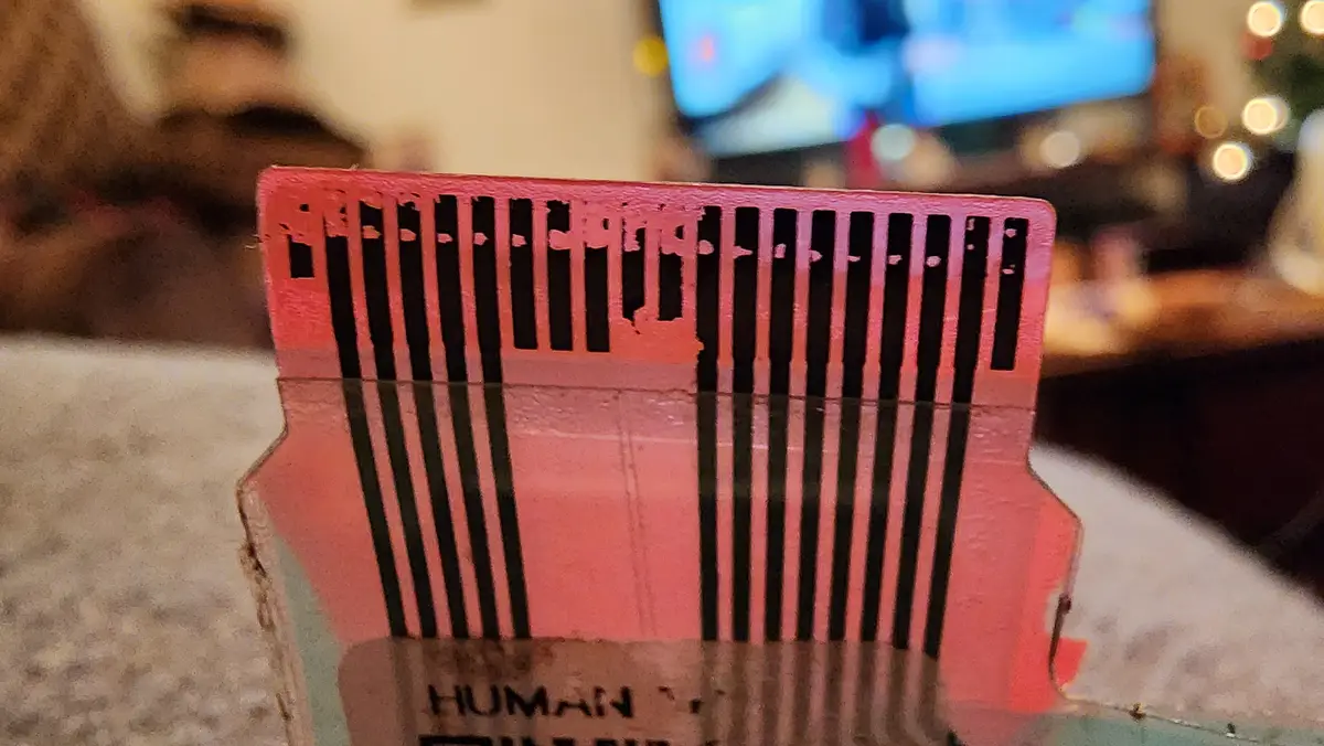 A close-up of a pink, 3D-printed object resembling a barcode or comb with vertical black slits, sitting on a textured…