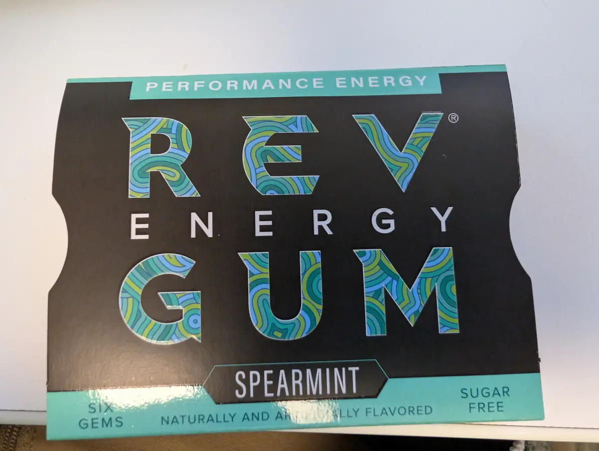 A close-up of a black and teal package of Rev Energy Gum, spearmint flavor, labeled 'Performance Energy' and 'Sugar Free'…