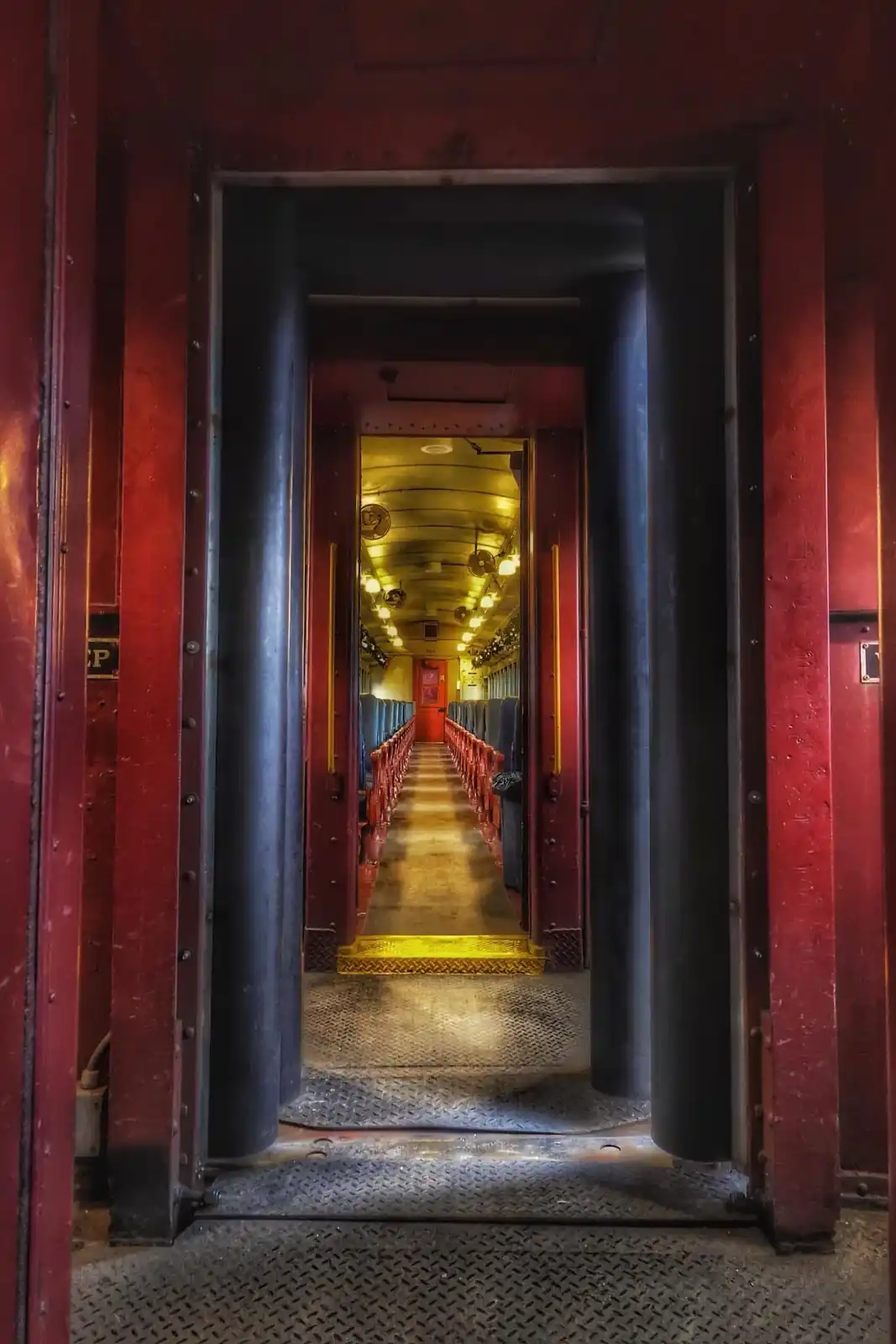 A view from the doorway of a vintage train car, looking down a long, empty aisle flanked by rows of blue seats and red trim…